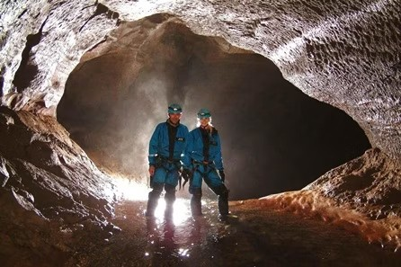 Two cavers in blue gear standing in a cave with an illuminated opening behind them.